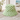 A green bucket hat with a white floral pattern, styled on a light wooden table with a vase of green leaves and a beach photo in the background.