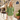 A woman stands on a city sidewalk wearing a green patterned blouse with rolled sleeves and light beige pants. The background shows a Parisian-style street scene with a cafe, potted plants, and a Rue Cler street sign.
