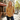 A woman stands on a Parisian street sidewalk wearing a yellow button-down shirt with a red and green floral pattern, paired with dark blue jeans. A cafe named 'Café de Flore' is visible in the background on the left.